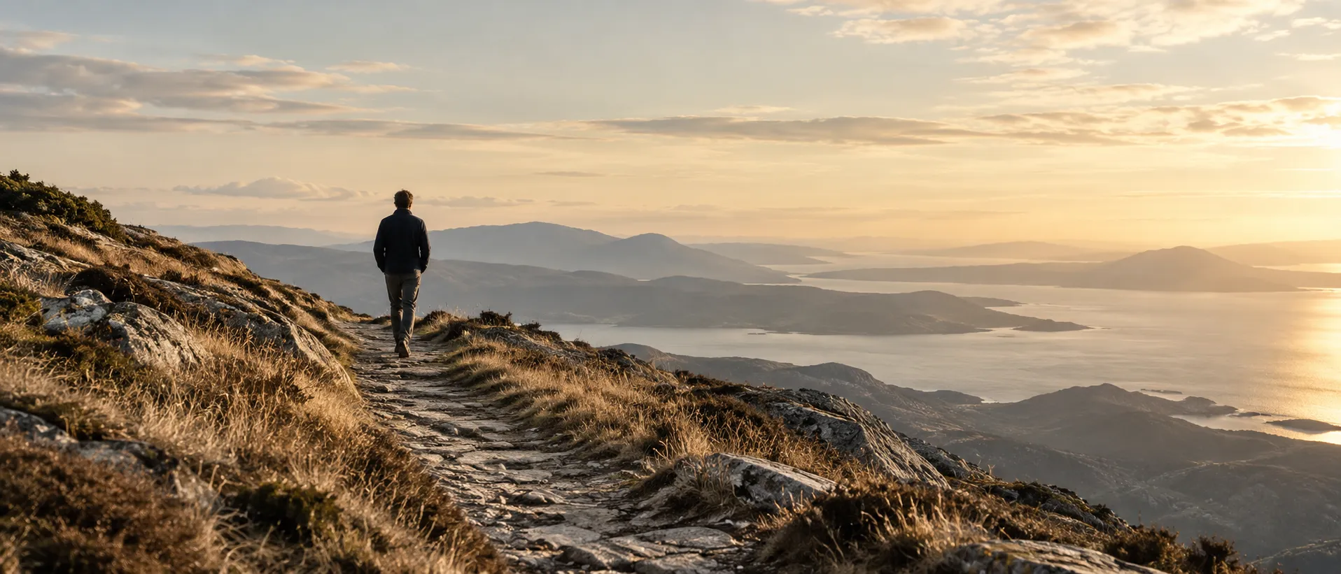 A person walking alone along a stone path across open highland at sunrise, the route ahead clear and unhurried.